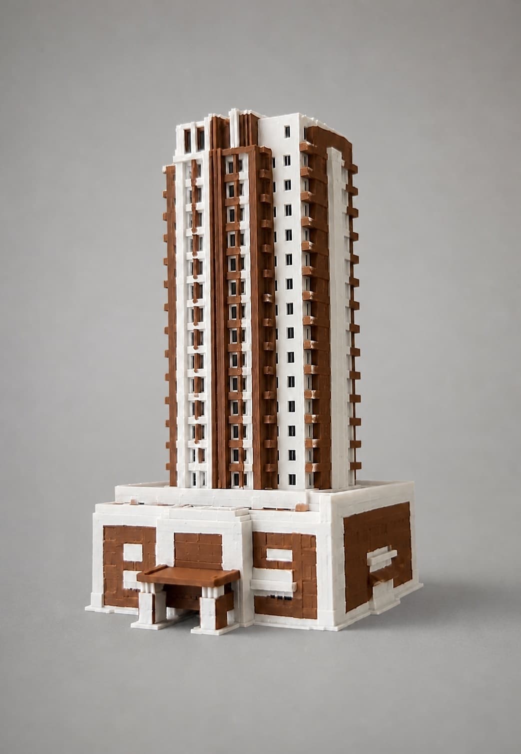 Painted brown-and-white apartment-building model standing alone on a neutral studio surface, showing the tower, podium and entrance at close range.